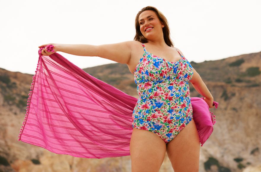 Woman with long brown hair wears a bright floral one piece swimsuit and holds a bright pink cotton sarong
