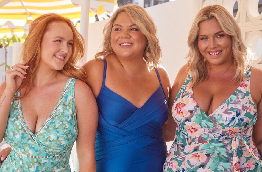 Three women stand closely together smiling. They each wear a different style of one piece swimsuit