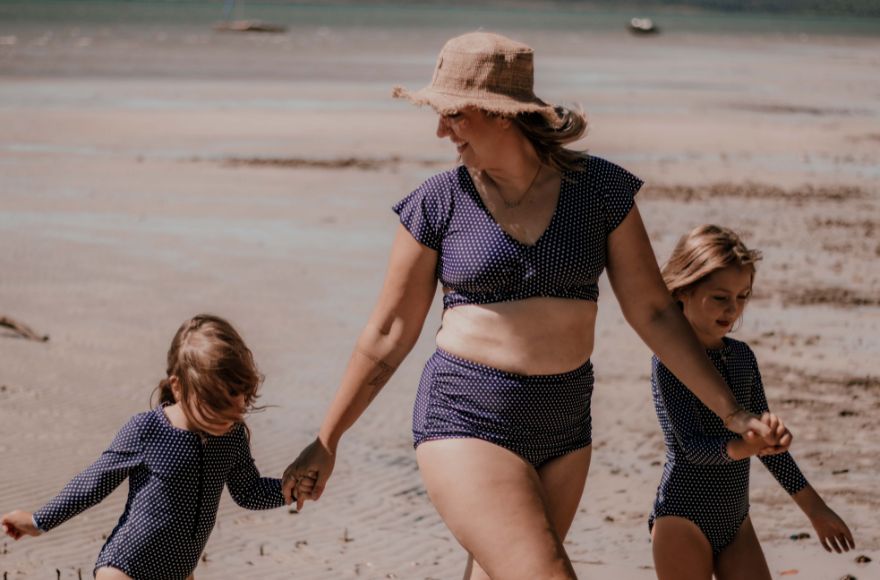 A woman dressed in Navy and White dots bikini set holds hands with her 2 daughters. They are walking on the beacher