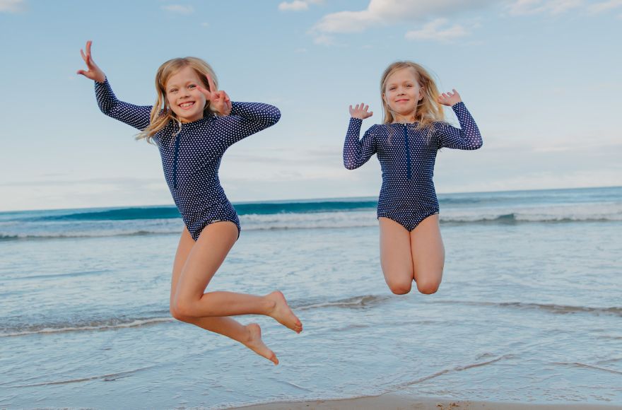 2 girls jump for joy at the beach wearing matching navy and white polkadot swimsuits. 