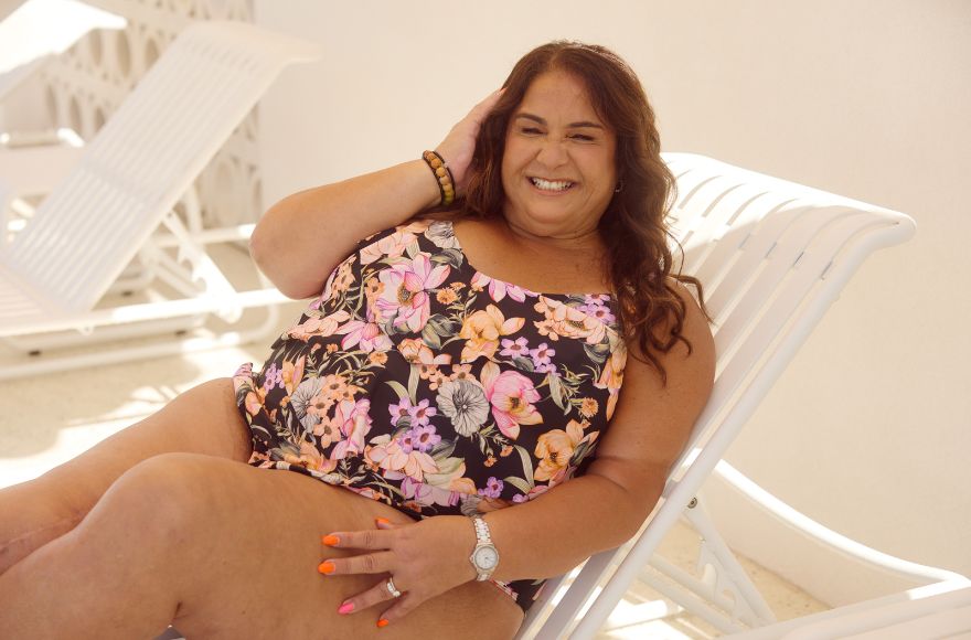 Woman with long brown hair reclines on a white sun lounge wearing a black and floral one piece swimsuit