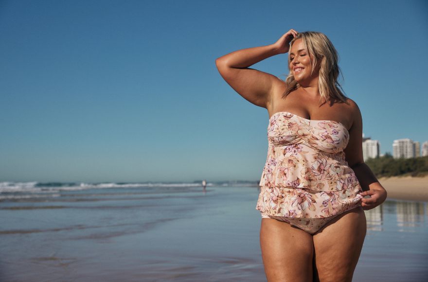 Woman with blonde hair poses on the beach wearing a soft pink floral strapless, tiered tankini top.
