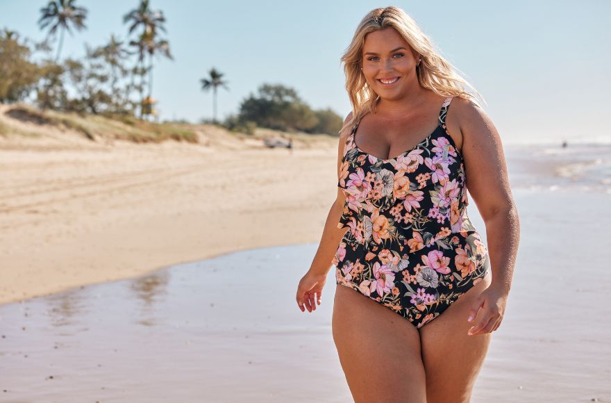 Woman with blonde hair walks along the beach wearing black swimsuit with pink and orange flowers