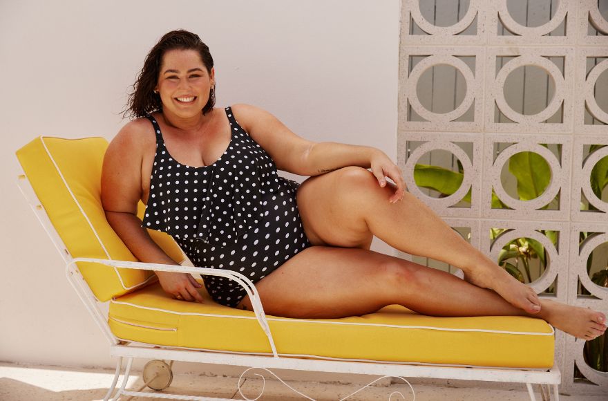 Woman reclines on yellow sunlounger wearing black and white one piece swimsuit with a frill detail. 