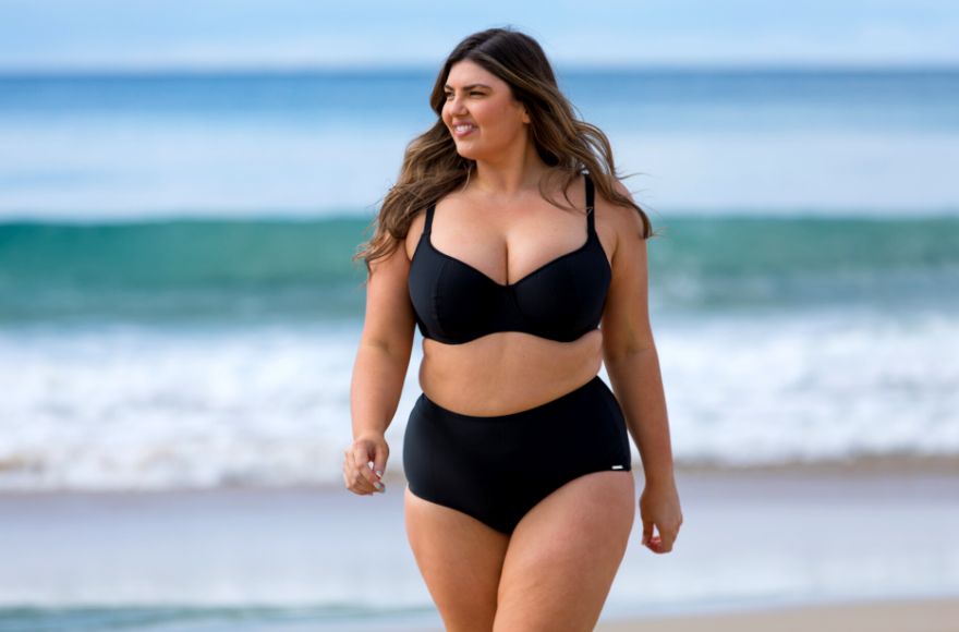 Woman with long brown hair walks on the beach wearing black underwire bikini top and high waisted pants