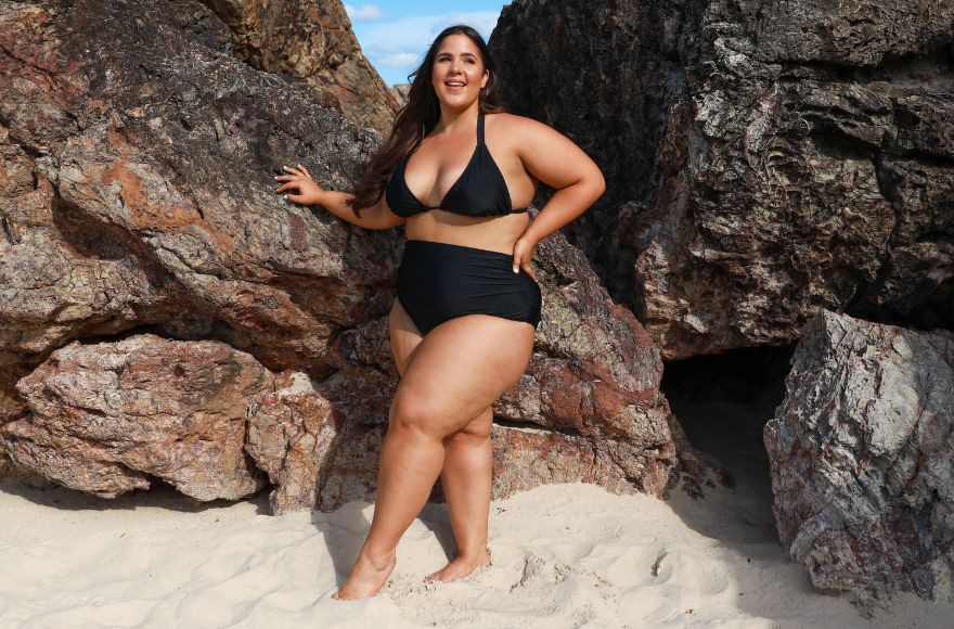 Woman with long brown hair poses at the beach wearing black triangle bikini and high waisted pants