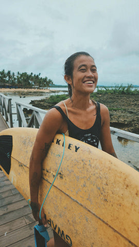 A smiling woman carrying a surfboard on a boardwalk built on a shallow reef.