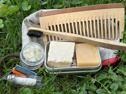 Kleiner Waschbeutel mit Holzzahnbürste, Holzkamm, Sheabutter, Zahnseide, zwei Seifen arrangiert im Gras.