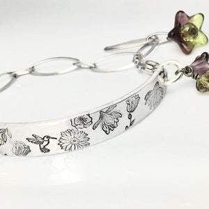 A silver bracelet with hummingbirds and flowers stamped on it. There are blue and purple beads dangling off the clasp.