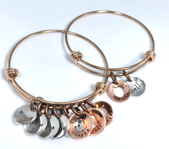 Two copper toned bangles are on a white background. Each bracelet has charms with hand stamped names on them.