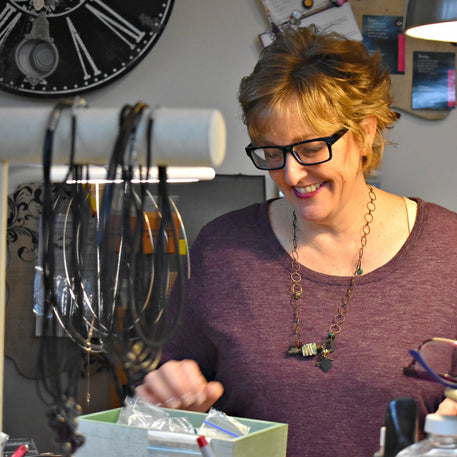 A woman in a purple shirt is looking down at a jewelry workbench.