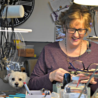 A woman in a purple shirt is looking down and using a tool to make jewelry. I little white dog is sitting next to her.