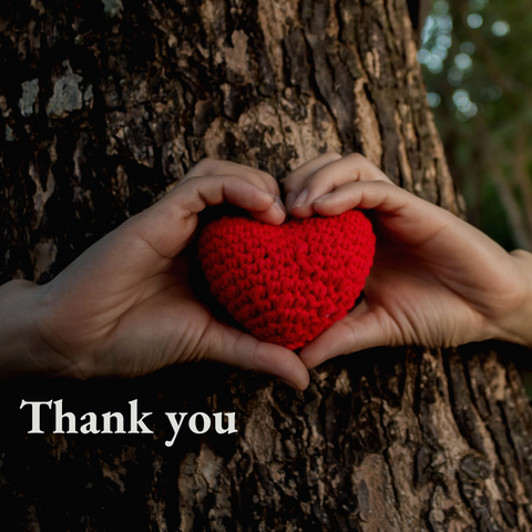Red knitted heart held between two hands forming the shape of a heart against a tree with the words saying thank you.