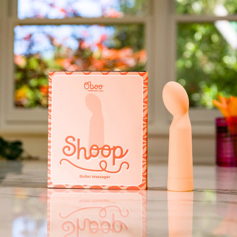 Peach-colored bullet massager placed next to its “Shoop” packaging by Oboo, set on a glossy surface with window light.