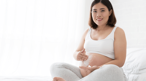 Pregnant Asian woman applying whipped shea butter, jojoba oil, and rosehip oil stretch mark cream to belly during pregnancy.