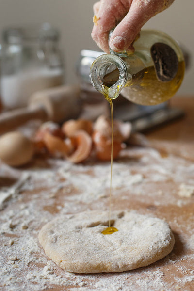 A person pouring fresh olive oil from glass jug onto rolled pizza dough