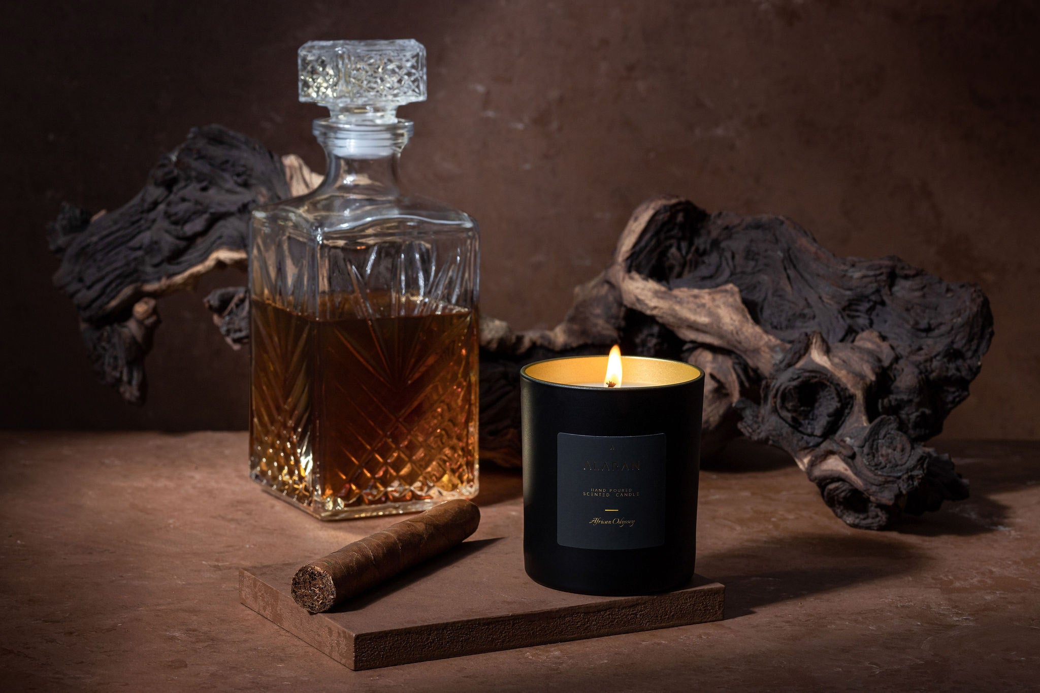 African Odyssey lit candle next to a cigar and crystal cognac decanter on a brown background