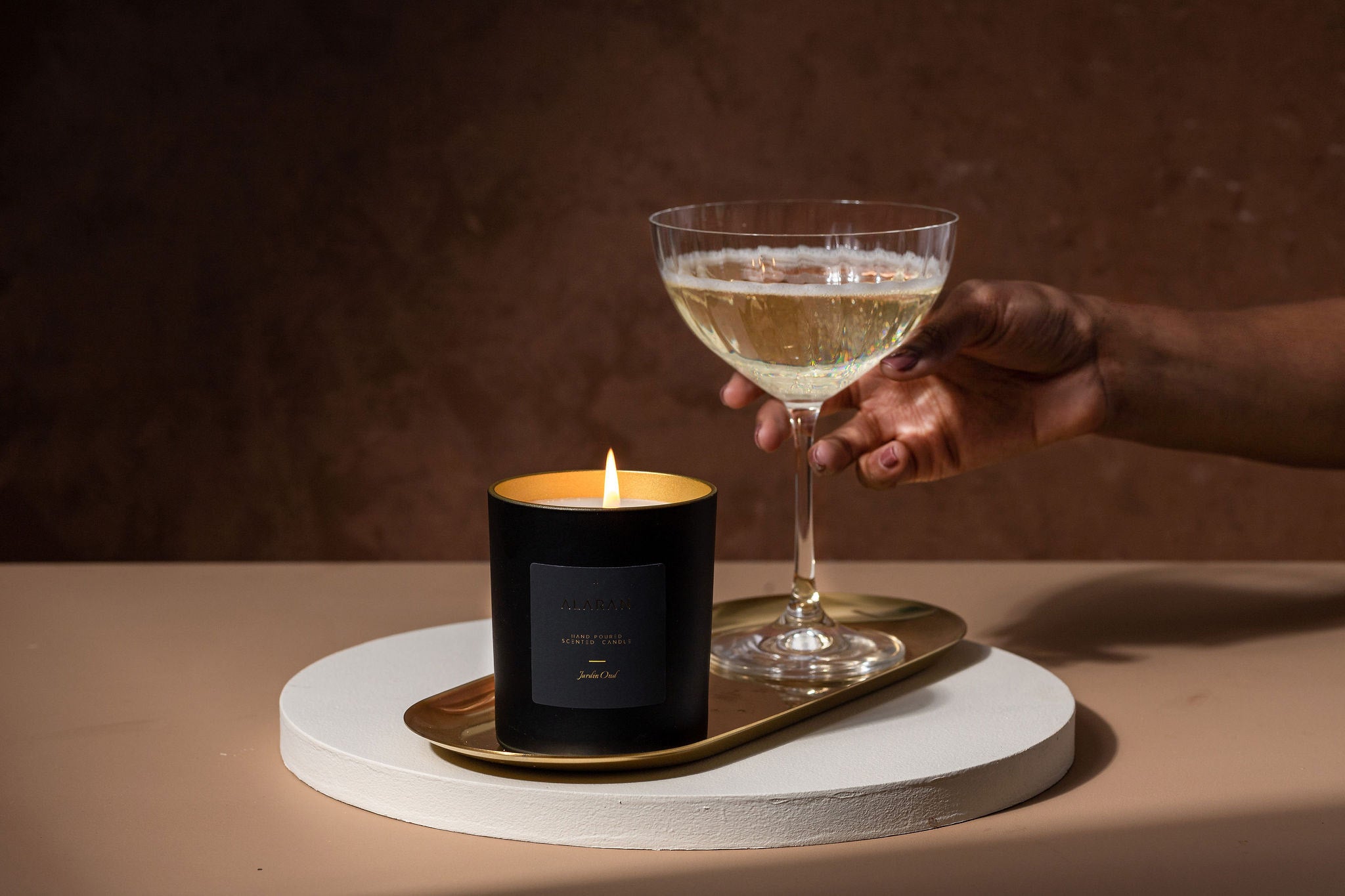 Jardin Oud lit candle on a gold tray next to champagne glass being picked up by a hand on a brown background