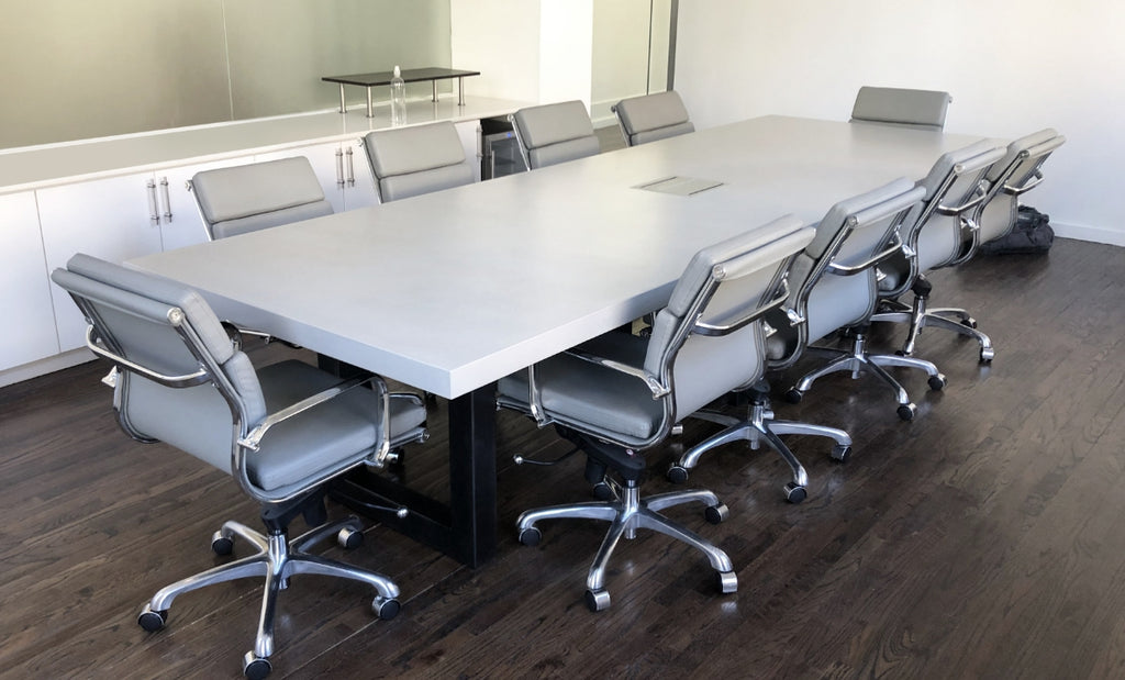 Conference table in light concrete surrounded by gray office chairs on top of a dark wood floor.
