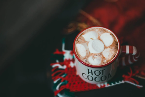 A white and red ceramic mug with hot chocolate garnished with 5 marshmallows, on a red and white woollen scarf