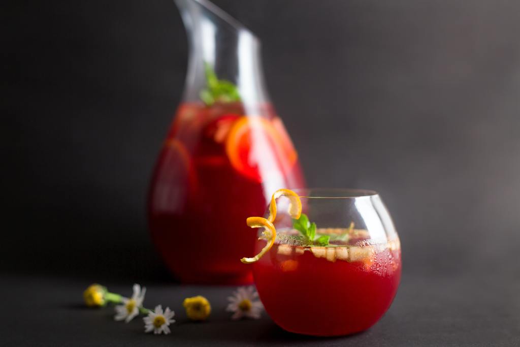 Small, round glass of red coloured alcohol-free Sangria with fruit decoration with a tall glass jug of alcohol free Sangria behind set against a dark grey background