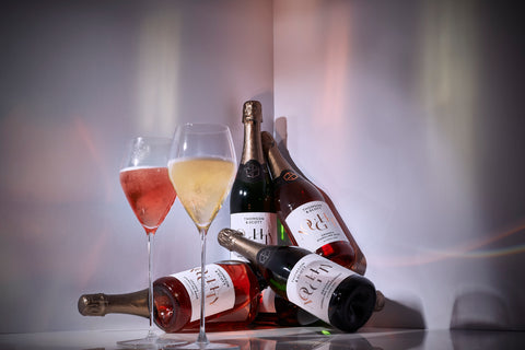 Two bottles of Noughty alcohol-free Chardonnay and two of Noughty alcohol-free rosé displayed in a corner against a white surface and background with a glass of each in wine glasses in the foreground