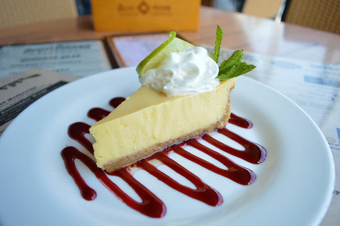 Slice of lemon tart displayed on a white plate with red sauce drizzled in a zigzag with white cream dolloped on top of the tart