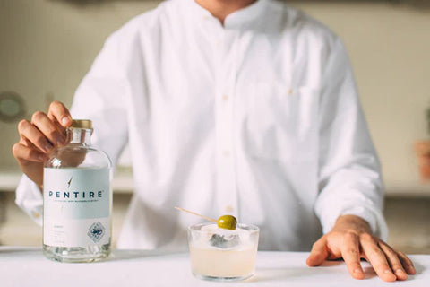 Person in white chef's jacket standing behind a white table with their hand on a bottle of Alcohol-free Pentire Adrift and an alcohol-free Pentire Adrift dirty martini also on the table