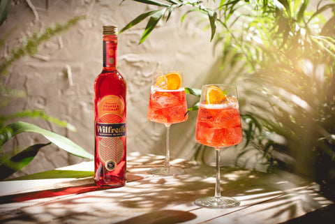 Bottle of Wilfred's non-alcoholic aperitif displayed next to two glasses of Wilfred's cocktails displayed against a white brick wall with plants around