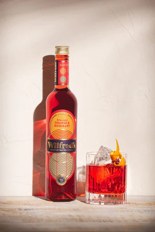 Bottle of Wilfred's alcohol-free aperitif red in colour with sunset coloured label pictured next to a glass of Wildred's Zero Proof Negroni cocktail garnished with an orange tweel