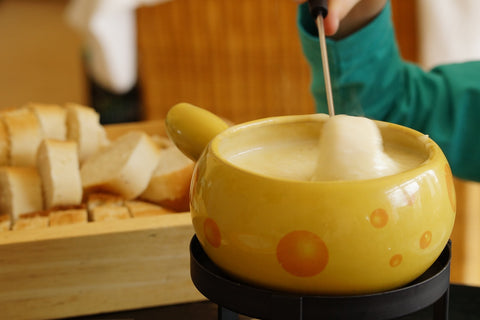 Cheese fondue in a yellow bowl with someone wearing a green sleeved top with only the arm visible dipping in a piece of bread and slices of bread on a wooden board in the background