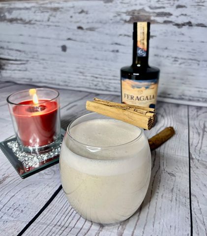 Alcohol free vegan eggnog cocktail displayed on white wooden background with candle and bottle of Feragaia in background