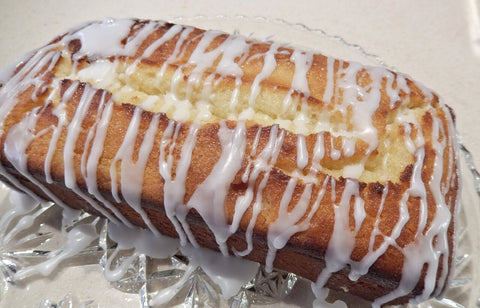 Lemon drizzle loaf cake with white icing drizzled over displayed on a glass plate with a white surface below and shot taken from above