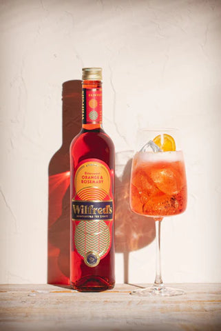 Bottle of Wilfred's alcohol-free Aperitif red in colour with sunset coloured label displayed next to a wine glass containing an alcohol free spritz by Wilfred's cocktail orange in colour and garnished with a slice of orange