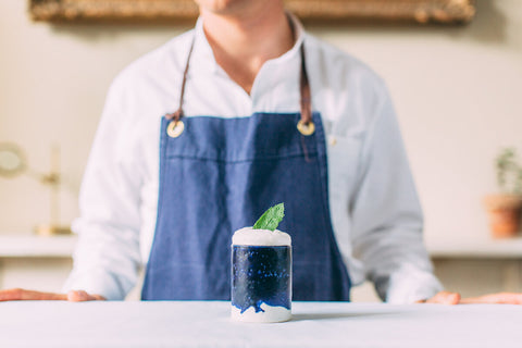 Male in a white shirt wearing a blue apron pictured from the chin down to the elbows behind a white table on which is a glass of Pentire adrift alcohol free oat fizz garnished with a mint leaf