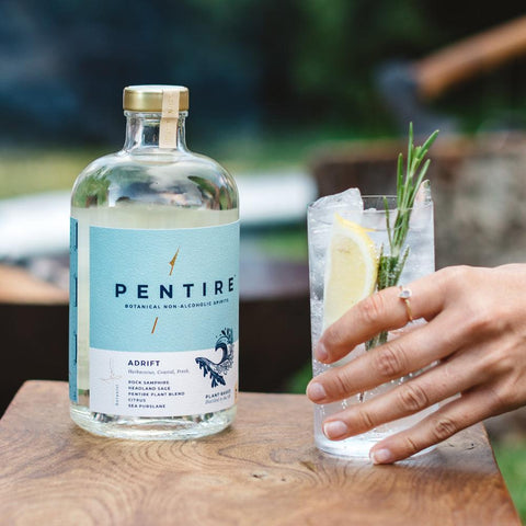 Bottle of Pentire Adrift displayed on a wooden table beside a glass containing Pentire Adrift and tonic held by a female hand with rosemary and lemon slice garnish