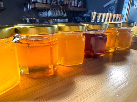 Six small hexagonal jars of honey of varying colours, set on a wood counter.