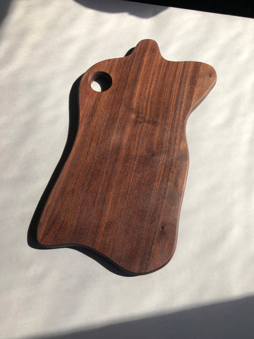 Photo of a cutting board made out of walnut. The shape of the board is an organic wavy shape. There's a finger sized hole on the top left of the board.