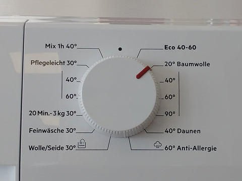 Programmwahlknopf einer Waschmaschine. Die Programme sind rund um den Knopf angeschrieben. Auf dem Knopf befindet sich ein erhabener Steg, der sich in Rot stark vom weißen Untergrund abhebt.