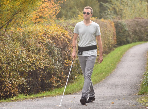 Blinder Mann geht auf einem Fußweg im Grünen. Um den Bauch trägt er den Navigürtel von Feelspace. In der Hand hält er einen Blindenstock.