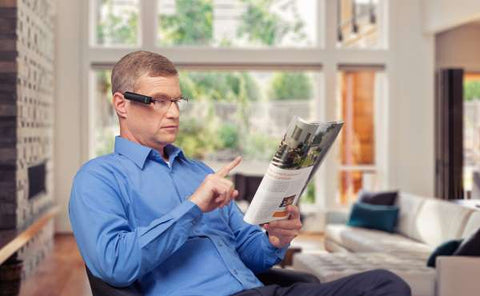 Ein Mann sitzt in einem Sessel und schaut in eine Zeitschrift. An seiner Brille ist eine Orcam befestigt.