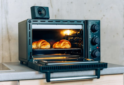 Ein Minibackofen mit Sprachausgabe. Die Tür des Backofens ist nach vorne aufgeklappt. Im Garraum befinden sich zwei goldbraune Croissants. Auf dem Backofen steht der Lautsprecher der Sprachausgabe.