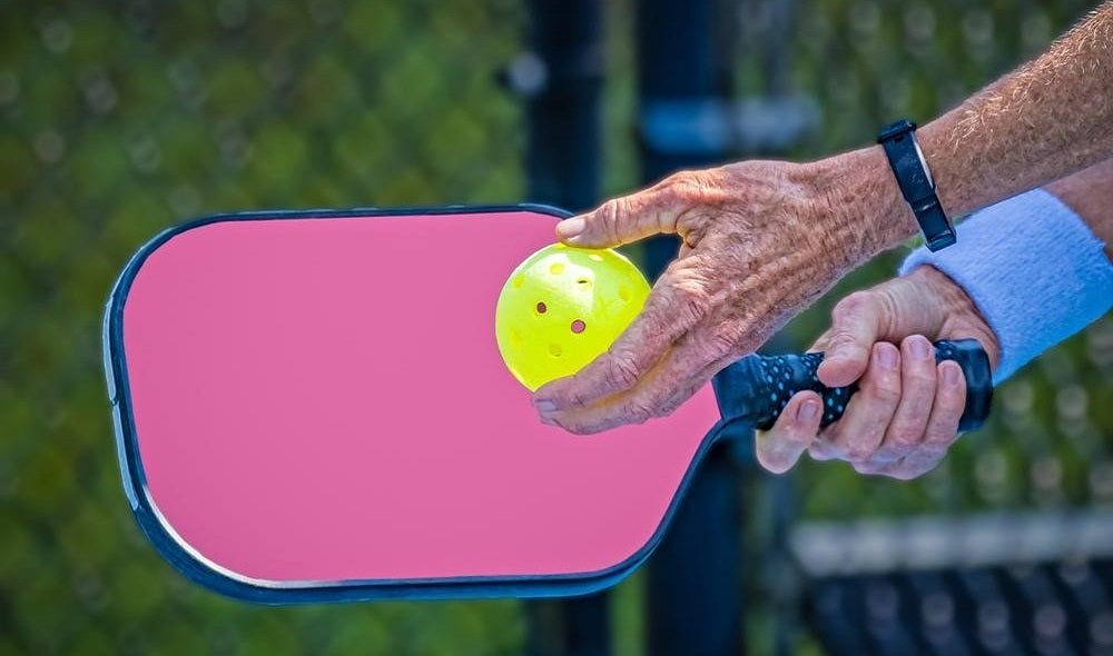 old hands with pickleball paddle and yellow ball