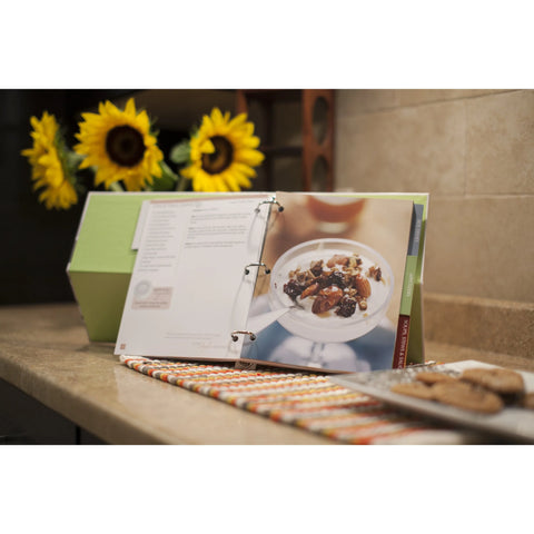 Open cookbook displaying a dessert recipe with a bowl of mixed ingredients, placed on a kitchen counter with sunflowers in the background.