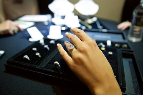 Photo of hand trying on large diamond pave engagement ring with many trays of rings below hand.