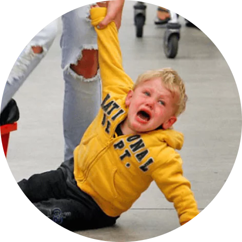 A crying child in a yellow hoodie being held by the arm in a public place.