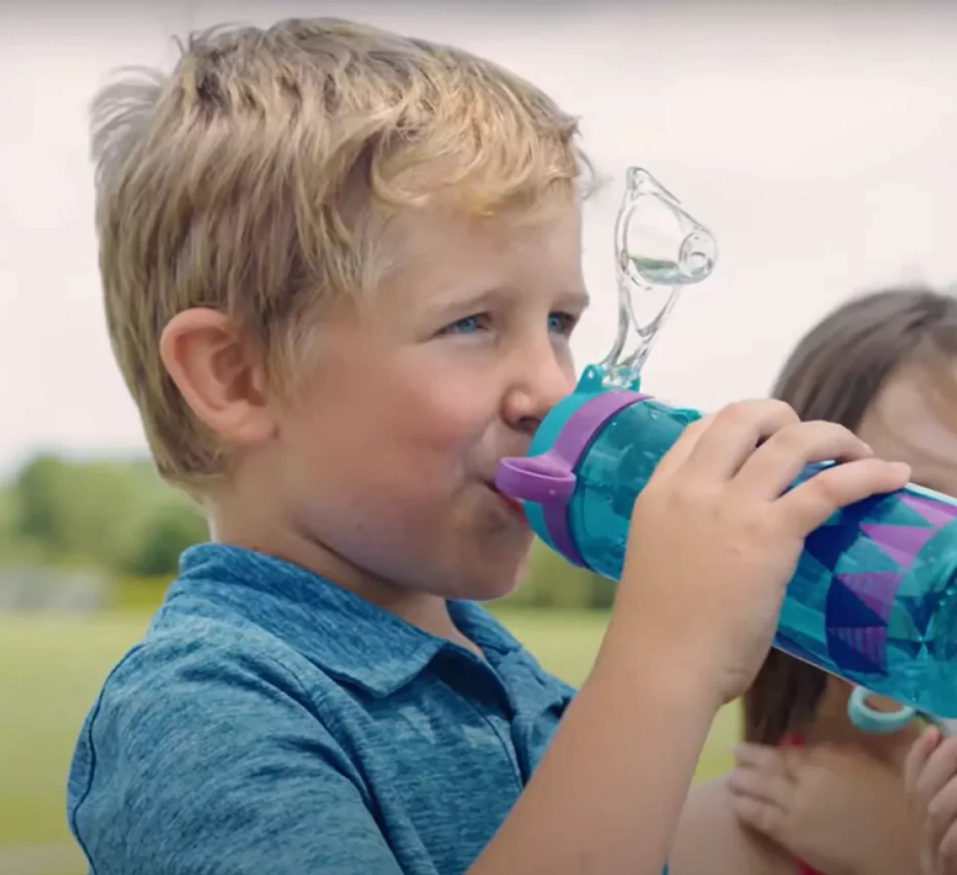 Child drinking from a blue water bottle with a straw outdoors.
