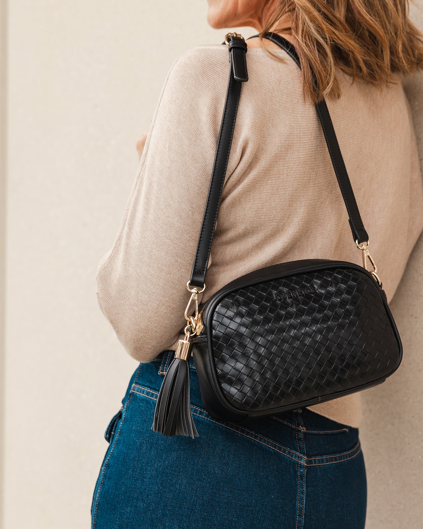 A woman wearing a beige knit sweater, holding a black woven crossbody bag over her shoulder