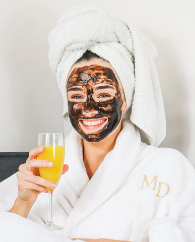 Woman in a dressing robe holding a peach Bellini and smiling while she has a face mask on. 