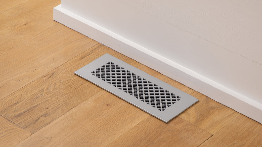 Silver metal vent cover in a light wood floor against a white wall.